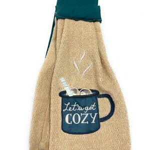 Pu&ograve; includere: Asciugamano da cucina beige con una tazza ricamata in blu e bianco che dice "Let's get cozy". L'asciugamano ha un'ansa di sospensione verde.