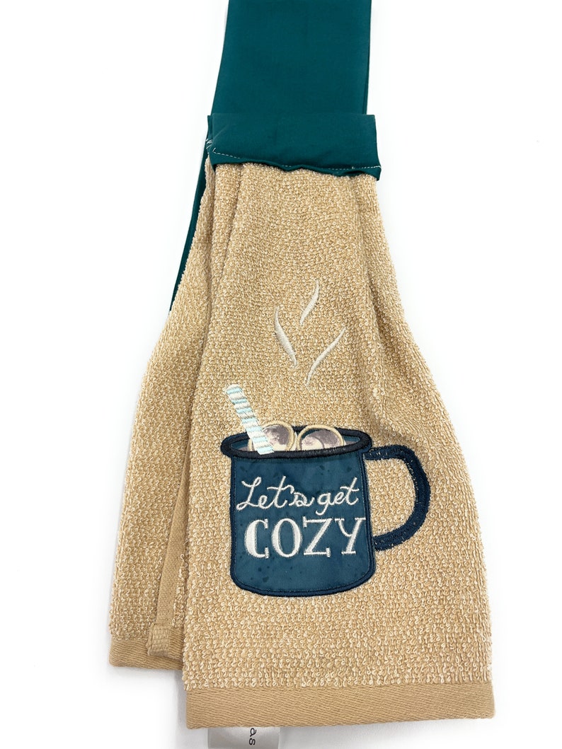 Pu&ograve; includere: Asciugamano da cucina beige con una tazza ricamata in blu e bianco che dice "Let's get cozy". L'asciugamano ha un'ansa di sospensione verde.