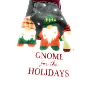 Pu&ograve; includere: Un asciugamano da cucina grigio e bianco con tre gnomi che indossano cappelli rossi, verdi e a quadri. L'asciugamano ha il testo "Gnome for the Holidays" ricamato.