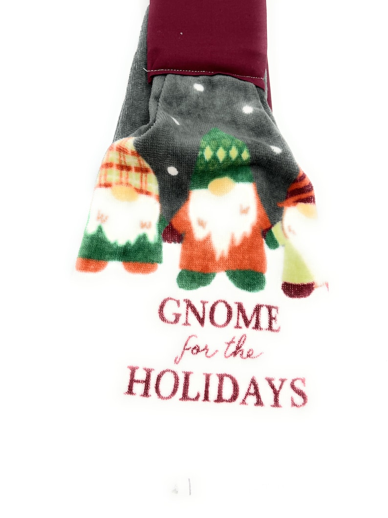 Pu&ograve; includere: Un asciugamano da cucina grigio e bianco con tre gnomi che indossano cappelli rossi, verdi e a quadri. L'asciugamano ha il testo "Gnome for the Holidays" ricamato.