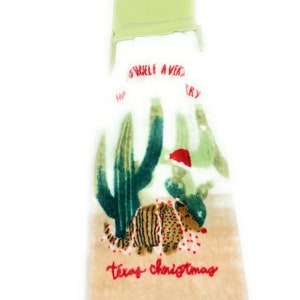 Pu&ograve; includere: Un asciugamano da cucina beige con un cactus verde e un armadillo rosso e bianco con un cappello da Babbo Natale. Il testo "Texas Christmas" &egrave; ricamato in rosso sull'asciugamano.