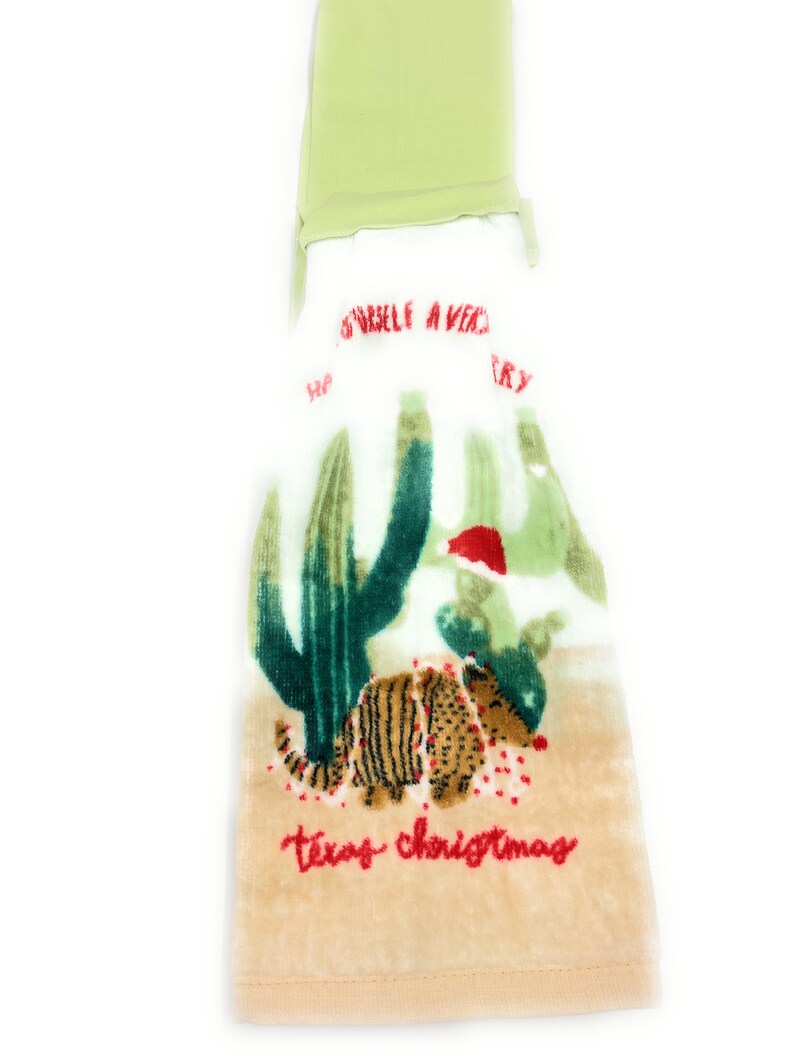 Pu&ograve; includere: Un asciugamano da cucina beige con un cactus verde e un armadillo rosso e bianco con un cappello da Babbo Natale. Il testo "Texas Christmas" &egrave; ricamato in rosso sull'asciugamano.