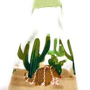 Pu&ograve; includere: Un asciugamano da cucina bianco con un armadillo marrone che indossa una collana di perline rosse e bianche. L'armadillo &egrave; circondato da cactus verdi su uno sfondo marrone.