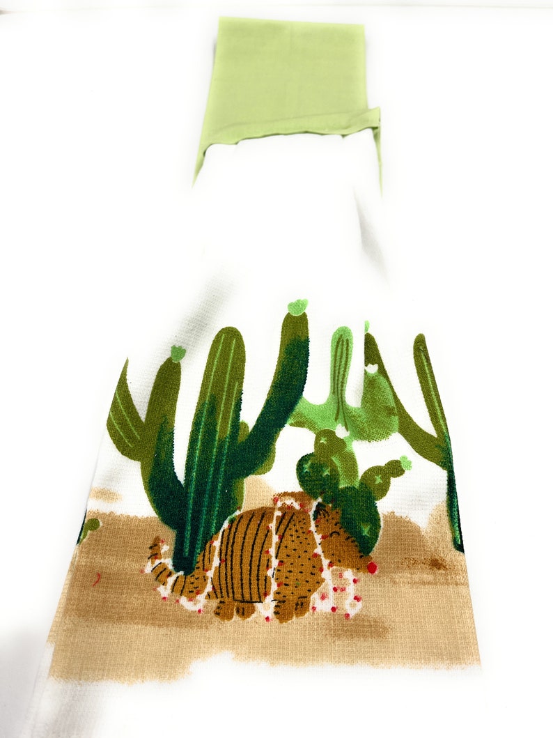 Pu&ograve; includere: Un asciugamano da cucina bianco con un armadillo marrone che indossa una collana di perline rosse e bianche. L'armadillo &egrave; circondato da cactus verdi su uno sfondo marrone.