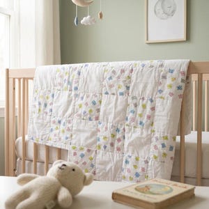 May include: A white patchwork baby blanket featuring a repeating pattern of small, colorful teddy bear motifs in blue, pink, and yellow. The blanket is draped over a light-colored wooden crib, with a cream-colored knit teddy bear and a book nearby.