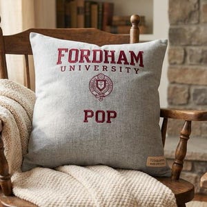 May include: A square light gray throw pillow featuring the maroon text Fordham University, a circular university seal, and the word Pop in maroon lettering. The pillow has a textured fabric finish and a small rectangular tag on the bottom right corner.