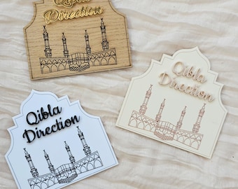 May include: Three arched signs featuring the text Qibla Direction in raised lettering. One sign is light wood with gold text, one is white with black text, and one is cream with light wood text. Each displays a line drawing of the Kaaba and minarets.