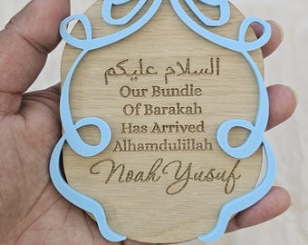 May include: A circular wooden plaque featuring a light blue acrylic bow frame. The plaque is engraved with the Arabic text السلام عليكم, followed by the English text Our Bundle Of Barakah Has Arrived Alhamdulillah, and the name Noah Yusuf in script.