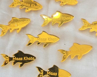 May include: A collection of gold-colored, reflective fish-shaped acrylic cutouts. Each piece features the engraved text "Maas Khata" and "S&K" on the surface. The fish are arranged in a scattered pattern, showcasing a smooth, metallic finish.