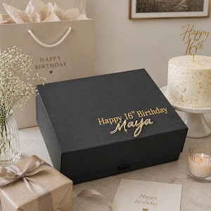 Pode incluir: Uma caixa de presente preta com a inscrição dourada "Happy 16th Birthday Maya" é o ponto focal. Um presente embrulhado, um bolo, uma sacola de compras e um cartão sugerem uma celebração de aniversário.