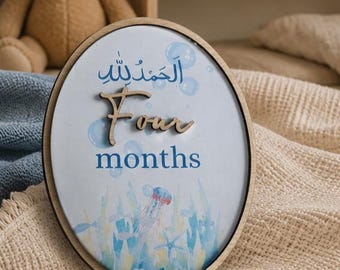 May include: An oval wooden plaque featuring a light blue background with watercolor illustrations of bubbles, seaweed, a jellyfish, and a starfish. The text reads "Alhamdulillah" in Arabic script, followed by the word "Four" in wood, and "months".