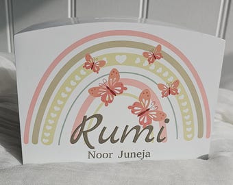 May include: A white arched wooden money box featuring a pastel rainbow graphic with small hearts and four orange butterflies. The name Rumi is printed in large dark script, with the name Noor Juneja printed in smaller dark text underneath.