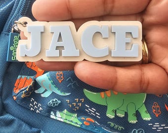 May include: A rectangular bag tag featuring the name JACE in raised light blue block letters on a cream-colored base. The tag is attached to a dark blue backpack decorated with a repeating pattern of green and orange cartoon dinosaurs.