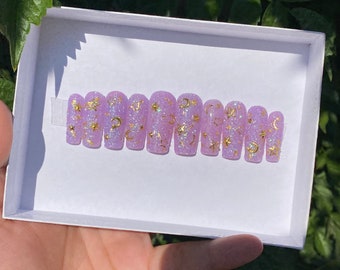 May include: A set of ten lavender-colored press-on nails with gold glitter and a design of stars and moons.