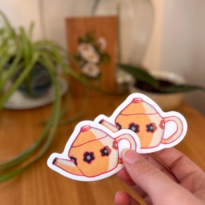 May include: Two stickers featuring a whimsical teapot design. The teapots are primarily orange with black flower accents and white handles and spouts. The stickers are held against a blurred background of a wooden surface and greenery.