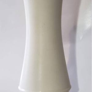 May include: A tall, off-white ceramic vase with a flared top and a ridged base. The vase has a smooth, curved body and stands on a white surface. The design is simple and elegant, suitable for various floral arrangements.