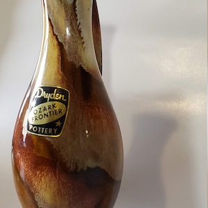 Könnte beinhalten: Eine Vintage-Vase von Dryden Ozark Frontier Pottery. Die Vase hat eine glänzende Oberfläche mit einer Mischung aus braunen, beigen und cremefarbenen Farben. Die Vase hat einen breiten Sockel und einen schlanken, gebogenen Hals. Das Dryden Ozark Frontier Pottery-Etikett ist sichtbar.