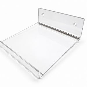 May include: A clear acrylic wall-mounted display shelf featuring a flat rectangular base, a front lip to prevent items from sliding off, and a vertical back panel with two pre-drilled circular holes for mounting hardware to a wall surface.