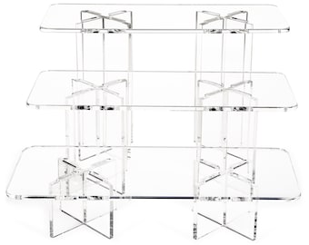 May include: Three-tier acrylic cake stand with clear rectangular shelves and a criss-cross base. The stand is made of clear acrylic and is perfect for displaying cakes, cupcakes, or other desserts.