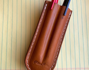 May include: A brown leather pen holder with white stitching. The holder has two slots for pens. A red pen and a black pen are inserted in the slots. The holder has the text "Palmer Leather" embossed on the front.