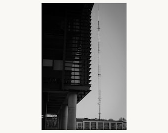 Op de afbeelding: Een zwart-wit architectonisch beeld van een modern gebouw met donkere horizontale lamellen aan de linkerkant, ondersteund door cilindrische kolommen, en een hoge, slanke metalen zendmast die op de achtergrond de lucht in steekt.