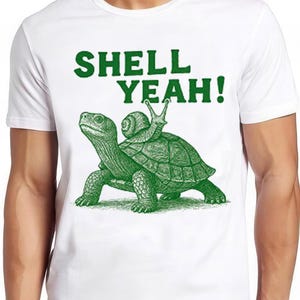 May include: White t-shirt featuring a green illustration of a turtle with a snail on its back, and the words "SHELL YEAH!". A fun design for everyday wear.