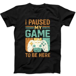 Op de afbeelding: Een zwart t-shirt met korte mouwen en een verweerde grafische print in oranje, groenblauw en wit. De tekst luidt I PAUSED MY GAME TO BE HERE, vergezeld van een centrale illustratie van een videogamecontroller met een versleten vintage textuur.