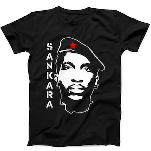 Op de afbeelding: Een zwart t-shirt met korte mouwen en een wit grafisch portret van een persoon die een baret draagt met een kleine rode ster. De tekst SANKARA is verticaal in witte hoofdletters aan de linkerkant van het portret op de voorkant gedrukt.