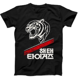 Puede incluir: Una camiseta negra de manga corta con un gráfico blanco de una cabeza de tigre rugiendo sobre una franja roja diagonal y texto en coreano. El diseño tiene una textura desgastada y está situado en el centro de la zona del pecho.