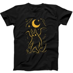 May include: A black short-sleeved t-shirt featuring a yellow graphic print of a winged creature standing beneath a crescent moon and stars. The illustration uses thin, textured lines to depict the creature, clouds, and celestial elements.