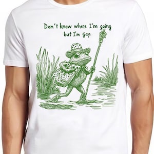 Peut inclure: T-shirt blanc avec une illustration verte d'une grenouille portant un chapeau et tenant un bâton. Le texte "Don't know where I'm going but I'm Gay." est imprimé sur le t-shirt.