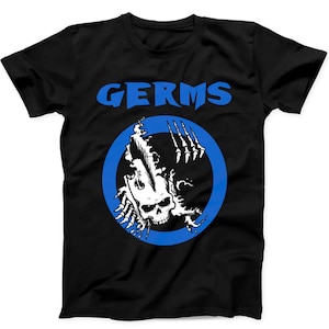 Könnte beinhalten: Schwarzes T-Shirt mit dem Wort "GERMS" in blauen Buchstaben über einem blauen Kreis. Im Kreis befindet sich ein weißer Totenkopf mit krallenartigen Händen und stacheligem Haar.