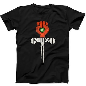 May include: A black short-sleeved t-shirt featuring a graphic print on the chest. The design displays a red fist clutching a green flower, positioned above a white dagger. The word GONZO is printed in white, distressed, block-style lettering.