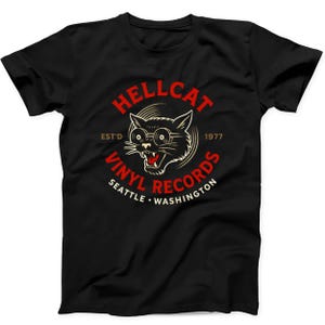 May include: A black short-sleeve t-shirt featuring a graphic of a cat wearing glasses with the text HELLCAT VINYL RECORDS EST'D 1977 SEATTLE WASHINGTON. The design uses red and cream colors to depict the cat and the surrounding circular text.
