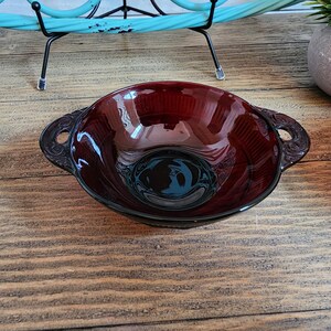 Può includere: Una ciotola di vetro rosso scuro con due manici. La ciotola ha una superficie liscia e lucida ed è leggermente curva. I manici sono sui lati della ciotola e sono anch'essi in vetro.