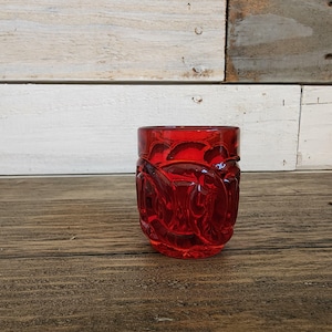 Puede incluir: Un vaso de chupito de vidrio rojo con un diseño decorativo. El vaso es transparente y tiene una superficie texturizada.