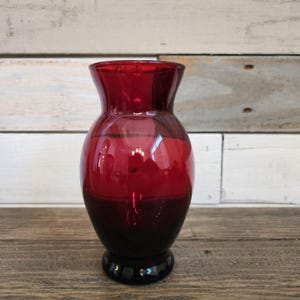 Peut inclure: Un petit vase en verre rouge avec un corps arrondi et un haut évasé. Le vase présente un dégradé de rouge foncé à noir.