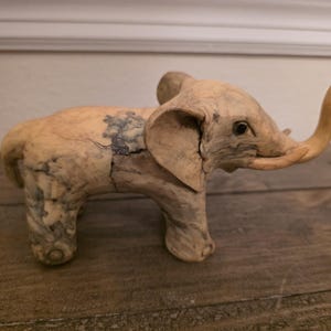 May include: A light-colored wooden elephant figurine with a raised trunk and large ears. The elephant has a textured surface with dark gray and black markings. The figurine is displayed on a wooden surface.