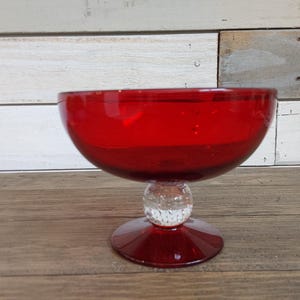 Puede incluir: Un cuenco de vidrio rojo con una base transparente y texturizada. El cuenco está sobre una superficie de madera.