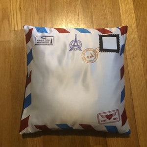 May include: A white pillow with a red, blue, and white striped border. The pillow has a printed design of an envelope with a stamp that says "Love Mail" and a stamp with a picture of the Eiffel Tower. The pillow also has a stamp that says "01-44-46 82 PARIS" and "REGISTRATION OFFICER".