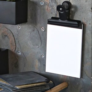 May include: A black metal clip holds a blank white notepad against a slate wall. The wall has a wooden shelf with a stack of black and brown paper on it.