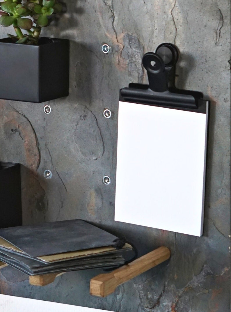 May include: A black metal clip holds a blank white notepad against a slate wall. The wall has a wooden shelf with a stack of black and brown paper on it.