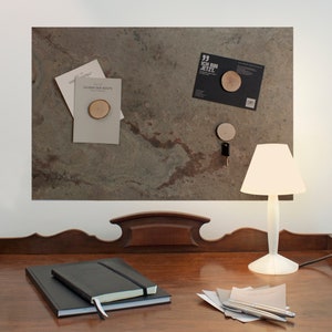 May include: A wooden desk with a dark wood finish. On the desk is a white lamp with a white shade. There are two black notebooks stacked on top of each other. There are also some papers and pens on the desk. The desk is in front of a wall with a brown and green slate tile surface. There are several items pinned to the slate tile surface, including a small wooden circle, a key, and a card with the text "Ich bin jetzt."