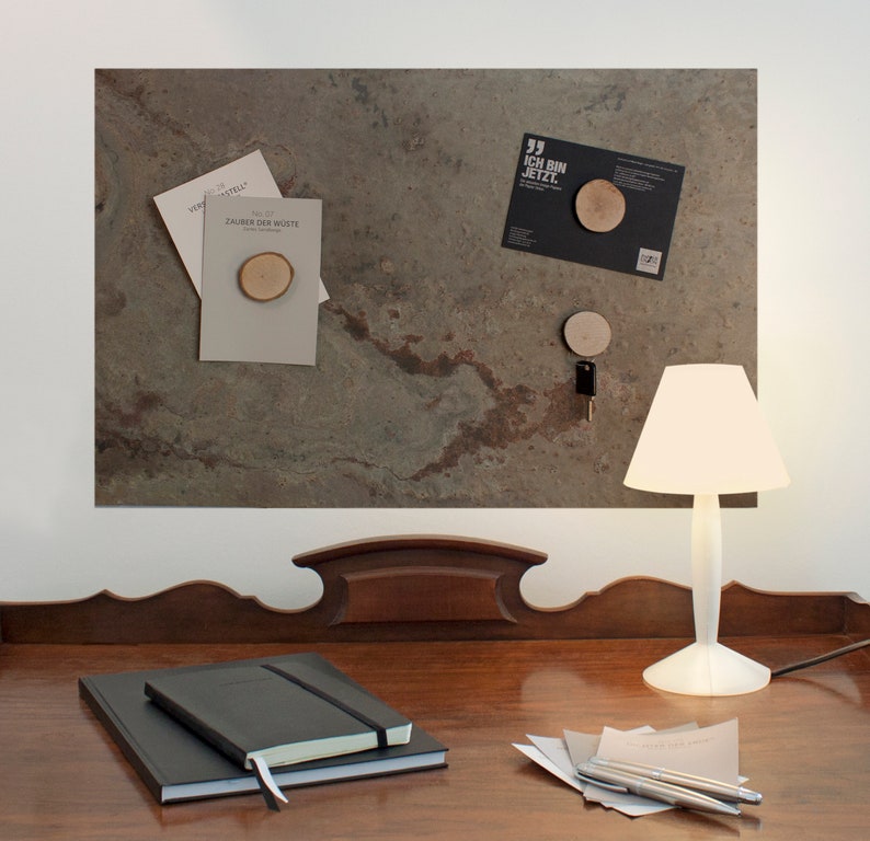 May include: A wooden desk with a dark wood finish. On the desk is a white lamp with a white shade. There are two black notebooks stacked on top of each other. There are also some papers and pens on the desk. The desk is in front of a wall with a brown and green slate tile surface. There are several items pinned to the slate tile surface, including a small wooden circle, a key, and a card with the text "Ich bin jetzt."