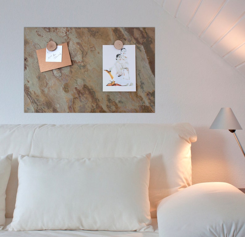 May include: A white headboard with two white pillows and a white throw pillow. A brown, textured, magnetic board with a photo and a note attached to it hangs on the wall above the headboard. A lamp with a white shade is on the right side of the bed.