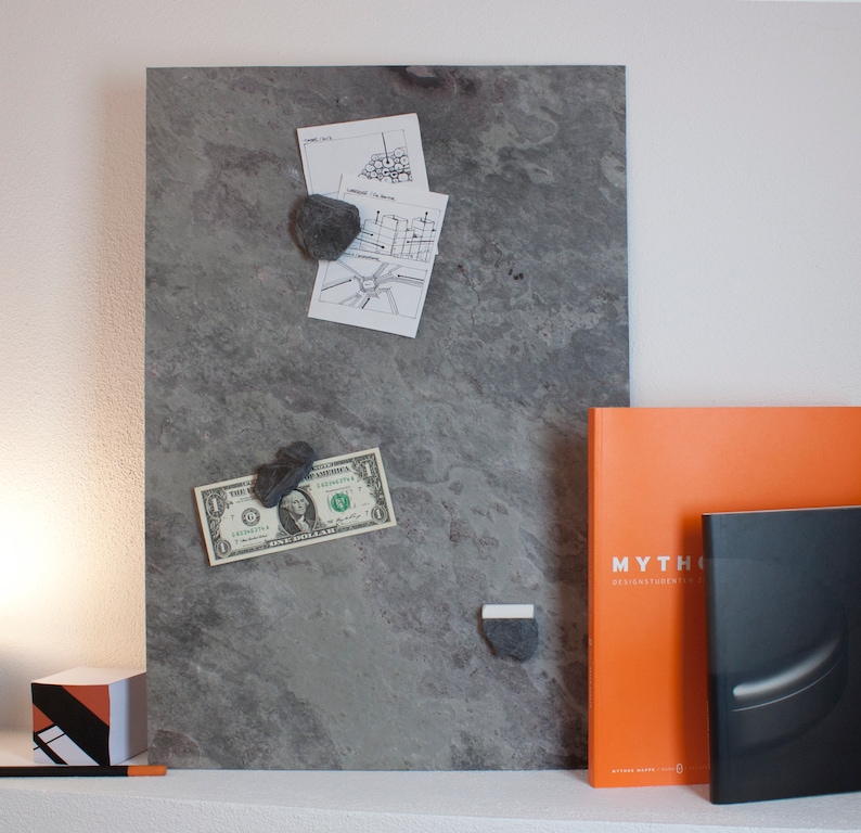 May include: A gray magnetic board with a dollar bill, a map, and a few small rocks attached to it with magnets. There are two books on a shelf below the board, one orange with the word "MYTHOS" on the cover and one black with a silver stripe.