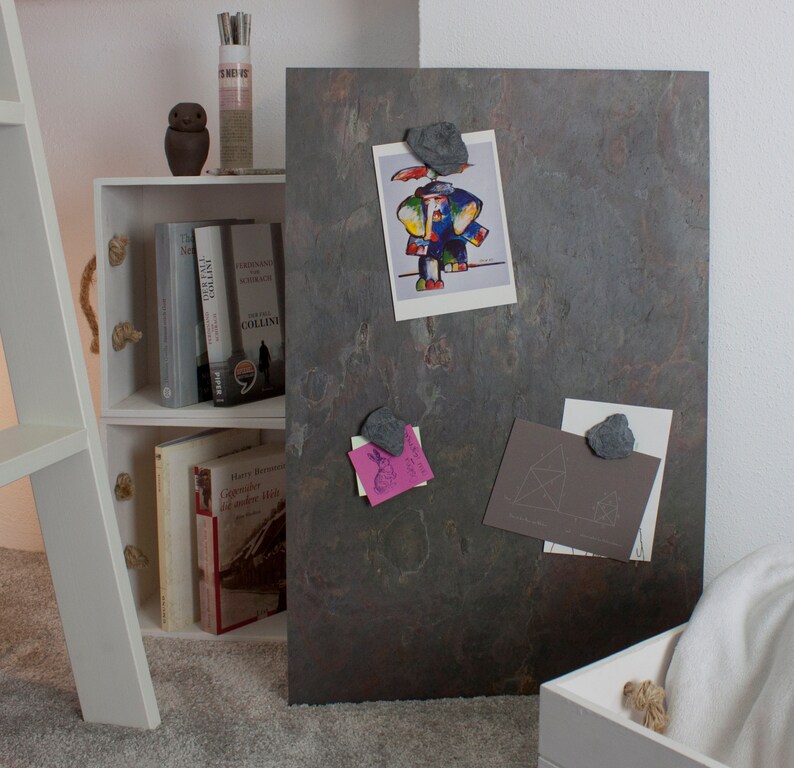 May include: A dark gray slate magnetic board with a variety of items pinned to it using small gray rocks. The items include a drawing of an elephant, a geometric design, and a pink note with a drawing of a bunny. The board is leaning against a white bookshelf with books on the shelves.