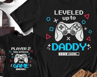 May include: Two black t-shirts with a video game theme. The larger shirt says "LEVELED up to DADDY" with a video game controller graphic and a loading bar. The smaller shirt says "PLAYER 2 has entered the GAME" with a video game controller graphic and a loading bar.