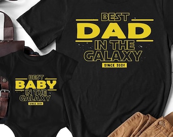 May include: Two black t-shirts with yellow text. The larger shirt says "Best Dad in the Galaxy Since 2024". The smaller shirt says "Best Baby in the Galaxy Since 2021".