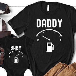 Könnte beinhalten: Zwei schwarze T-Shirts mit einer Tankanzeige und den Worten "Daddy" und "Baby". Die Anzeige ist unterschiedlich gefüllt, was darauf schließen lässt, dass das "Daddy"-Shirt fast leer und das "Baby"-Shirt voll ist.